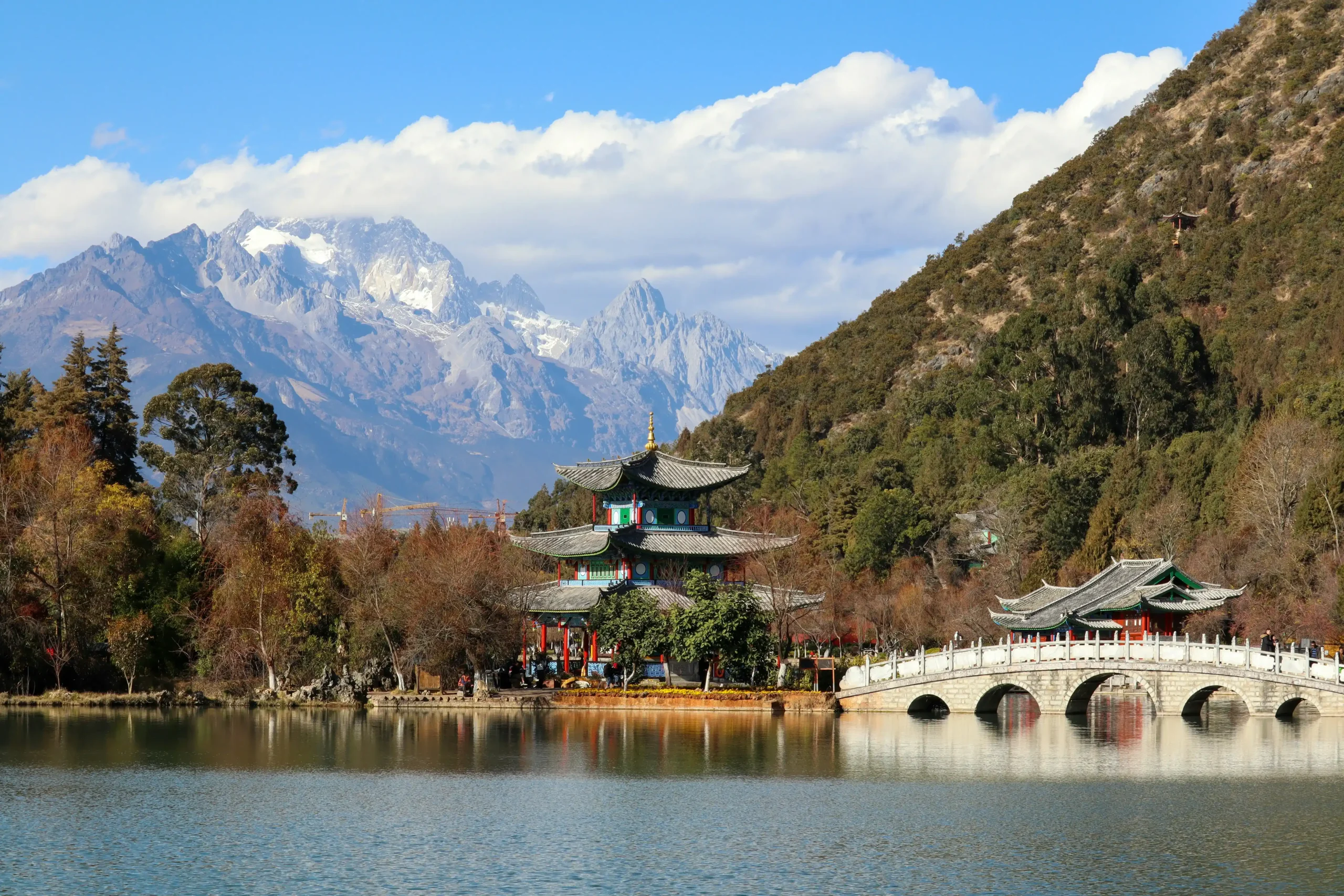 A traditional Chinese pavilion bridge arched over a calm pond, with Jade Dragon Snow Mountain visible in the distant background.