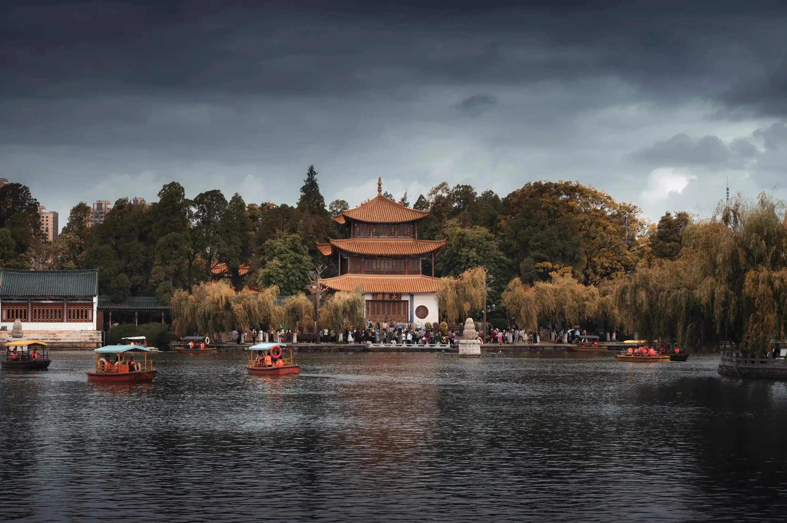 A historic multi-story Chinese pavilion surrounded by trees bordering a lake with boats floating on the water under a soft overcast sky.